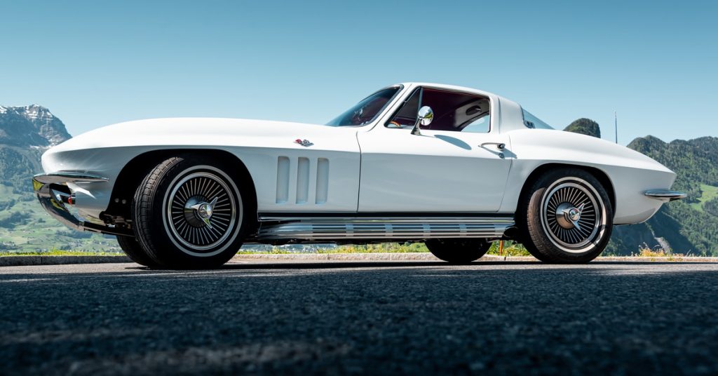 Low side view of a white classic sports coupe on asphalt, with mountains and clear blue sky behind it.